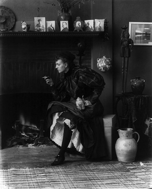 Frances_Benjamin_Johnston,_full-length_portrait,_seated_in_front_of_fireplace,_1896